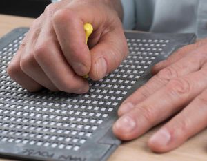 Oseba uporablja brajevo tablico in šilo