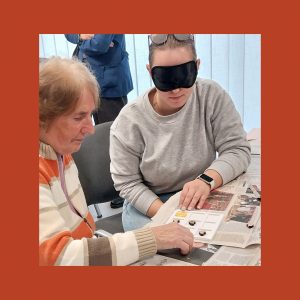 Na sliki sta 2 ženski, ena mlajša in ena starejša. Obe sedita za mizo. Mlajša ima na očeh temna očala, zaradi katerih ne more videti. Na mizi je časopisni papir. Na njem, med obema ženskama, pa je ročno izdelana igra: Tri v vrsto. Ženski igrata to igro.