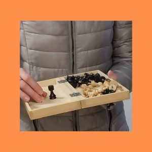 Na sliki je ženska v bež bundi. Vidi se njeno telo in roke, obraz in noge se ne vidijo. V rokah drži odprt šah, prilagojen slepim in slabovidnim.