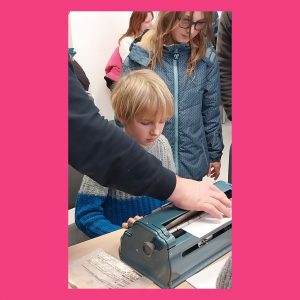 Na sliki sta 2 otroka, ki opazujeta brajev pisalni stroj. Vidi se tudi roka odraslega, ki bere kar je bilo na stroj napisano.