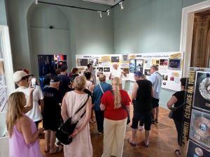 Na sliki je množica ljudi, obiskovalcev razstave v muzeju, oblečenih v poletna oblačila in večinoma obrnjenih s hrbtom proti nam. Poslušajo Andreja Šifrerja. ki stoji pred njimi in jim razlaga svojo zgodbo. Za Andrejem je velik pano, na katerem so pomembni mejniki v njegovem življenju,