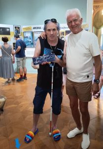 Na sliki sta Andrej Šifrer in slabovidni obiskovalec razstave, ki v rokah drži posvetilo avtorja. Andrej ima desno roko prijateljsko položeno na rame slabovidnega, oba sta obrnjena proti nam. V ozadju sta moški in ženska, ki si ogledujeta razstavo.