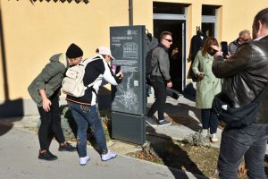 Dve osebi stojita pred informacijsko tablo z zemljevidom. Ena oseba s telefonom skenira QR kodo pri dnu table, druga ji pomaga pri usmerjanju. Okolica je mestna, pločnik in stavba sta v ozadju.