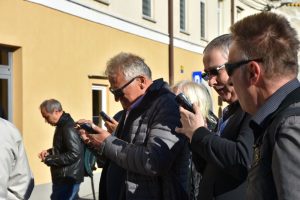 Skupina moških stoji na soncu in uporablja mobilne telefone. Videti je, da poslušajo zvočne posnetke ali berejo informacije na zaslonih. Nosijo temna očala in jakne, ozadje pa tvorijo rumena fasada in okna stavbe.