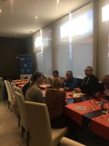 Skupina ljudi sedi okoli pravokotne mize v svetli sejni sobi. Na mizi so kozarci, vrč z vodo in papirji, udeleženci pa se pogovarjajo in sodelujejo pri skupinskem delu. V ozadju so velika senčena okna, pod njimi pa je videti promocijski pano z logotipi. Vzporedno ob mizi stojijo svetli tapecirani stoli.