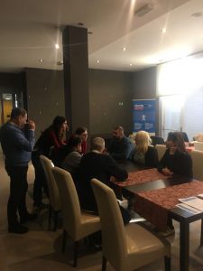 Večja skupina ljudi sedi in stoji okoli mize v hotelski sejni sobi. Udeleženci se pogovarjajo in delajo v manjših skupinah. V ozadju je moder pano z informacijami, na stenah pa majhne dekorativne lučke. Soba je nežno osvetljena, stolice so svetle barve, miza pa prekrita z rdečim prtom.