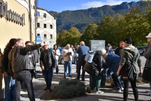 Skupina ljudi stoji na pločniku ob stavbi v sončnem dopoldnevu. Nekateri fotografirajo z mobilnimi telefoni, drugi poslušajo ali opazujejo okolico. V ozadju se dviga gorato zaledje, drevesa pa so v jesenskih barvah. V ospredju moški kaže proti nečemu izven kadra.