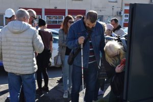 Večja skupina ljudi stoji na pločniku ob parkiranih avtomobilih. Moški z belo palico stoji v središču kadra, ob njem ženska uporablja telefon za skeniranje QR kode ali povečavo besedila. Ostali opazujejo ali pomagajo. Svetloba je močna, vidni so kontrasti senc.