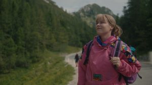 Na gozdni, gorski poti stoji oseba v rožnem pohodniškem anoraku in z nahrbtnikom. Oseba je obrnjena rahlo v desno, kot da opazuje pokrajino. Okoli nje je gosto zeleno drevje, v ozadju pa se dvigajo skalnati, poraščeni gorski vrhovi. Pot se vije skozi gozd in deluje kot priljubljena pohodniška trasa.