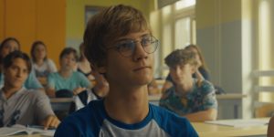 V šolski učilnici sedi skupina učencev. V ospredju je učenec v modri majici s kratkimi rokavi, ki gleda proti tabli ali učitelju. Za njim so drugi učenci, ki sedijo v klopeh, pred seboj imajo odprte zvezke in učbenike. Prostor je svetel, z rumenimi in svetlo sivimi stenami ter velikimi okni na desni strani, skozi katera prihaja dnevna svetloba.