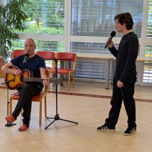 Dve osebi sredi nastopa. Levo sedi kitarist, ki poje v mikrofon, desno stoji Tilen Artač. Obrnjen je proti kitaristu, v roki ima mikrofon.