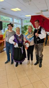 4 osebe stojijo, obrnjene so proti nam. Desno je moški v narodni noši, ima odprt dežnik. Levo od njega sta dve ženski v narodni noši, skrajno levo pa Luka Pavlin s klobukom na glavi.