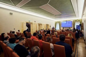 Udeleženci sedijo v konferenčni dvorani in spremljajo predstavitev projekta DATIS o dostopnem turizmu. V ospredju so vrste stolov z obiskovalci, v ozadju pa govorniški oder in projekcijsko platno.