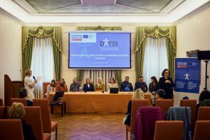 Okrogla miza v okviru projekta DATIS. Za mizo sedi več sodelujočih, ob strani stoji moderatorka z mikrofonom, nad njimi pa je projekcijsko platno z naslovom dogodka o dostopnem turizmu.