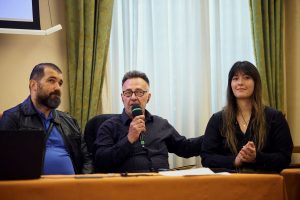 Trije udeleženci okrogle mize. Moški v sredini govori v mikrofon, ženska na desni ploska, slepi udeleženec na levi pozorno posluša.